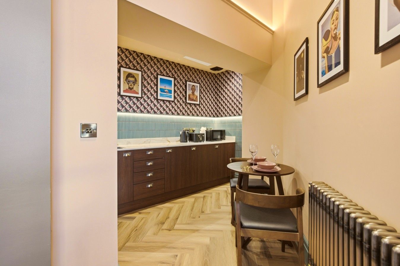 A compact kitchen with a herringbone floor pattern, wooden cabinets, a small dining table with chairs, and a radiator.