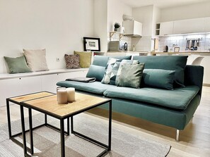 Ein modernes Wohnzimmer mit einem grünen Sofa, einem kleinen Holztisch und einer Küchenzeile im Hintergrund.
