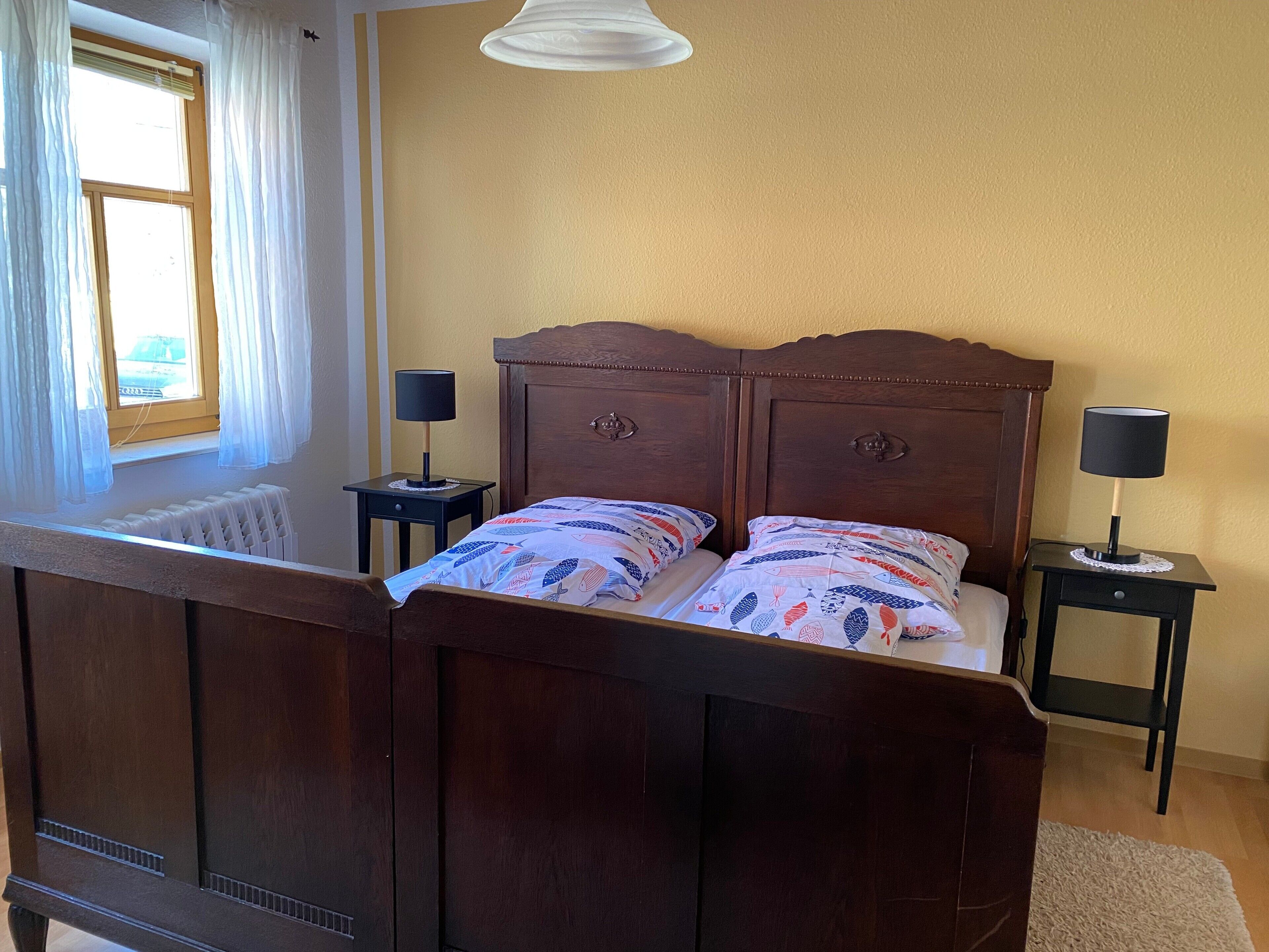 Una cama con cabecera, dos mesitas de noche con lámparas, una ventana con cortinas y un radiador.