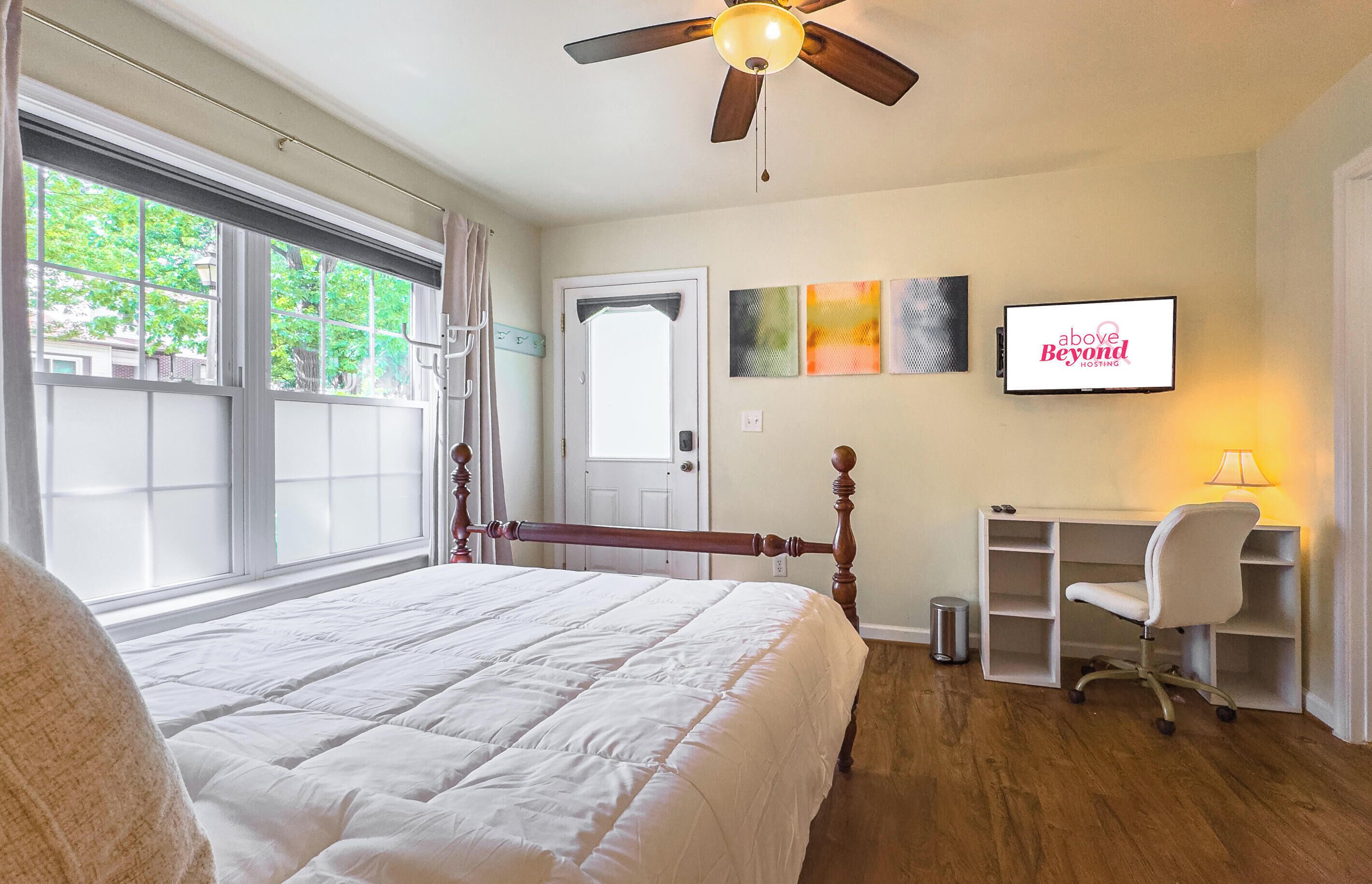 A bedroom with a bed, a desk, a chair, a ceiling fan, a television, and a window with curtains.
