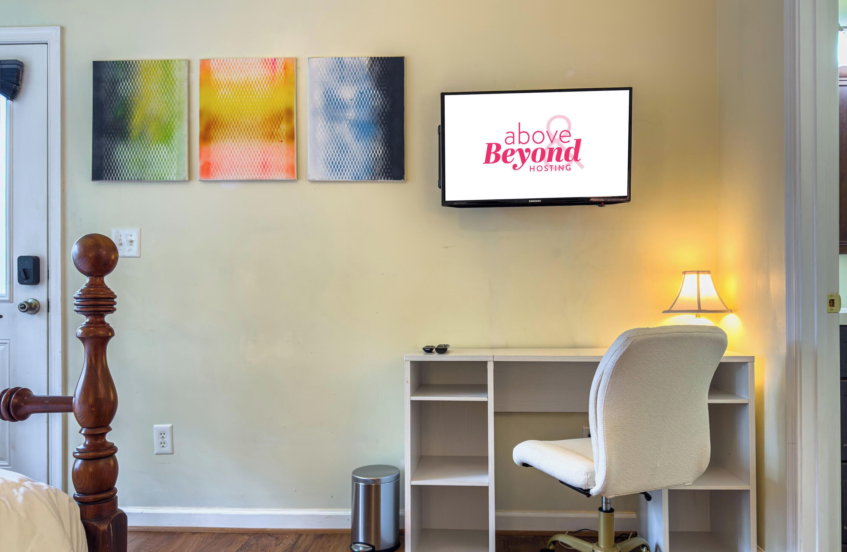 A hotel room with a bed, a desk with a chair, a TV displaying 'above Beyond Hosting,' and three abstract wall art pieces.