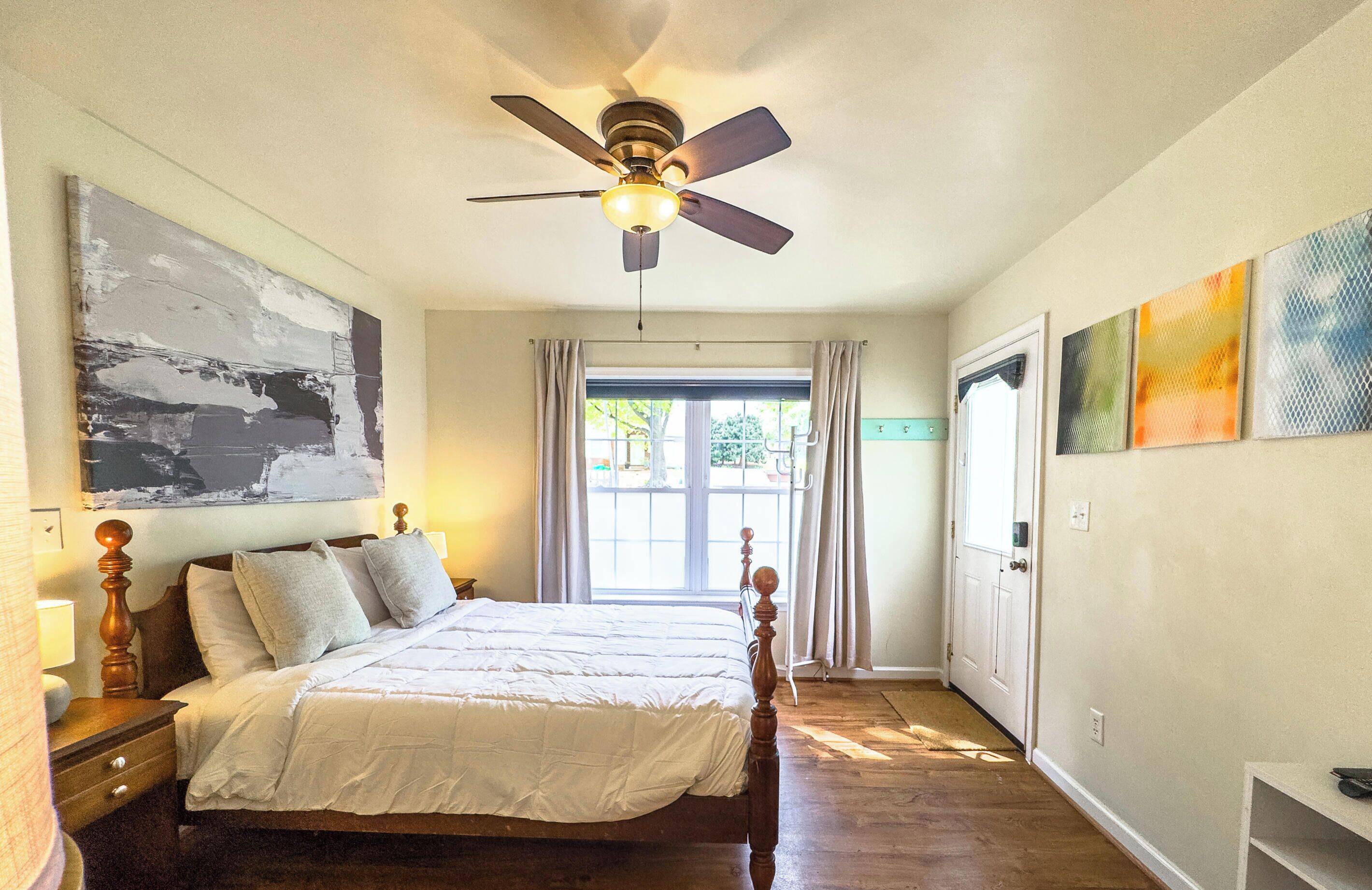 A bedroom with a bed, a ceiling fan, a window with curtains, and artwork on the walls.