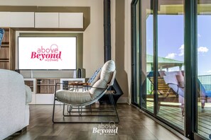 A modern living room with a large flat-screen TV displaying 'above Beyond HOSTING' logo, a comfortable lounge chair, and a view of a deck through floor-to-ceiling windows.