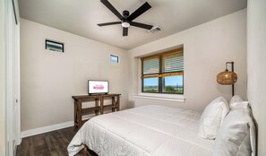 A bedroom with a bed, a ceiling fan, a window with blinds, a desk with a computer monitor, and a wall-mounted lamp.