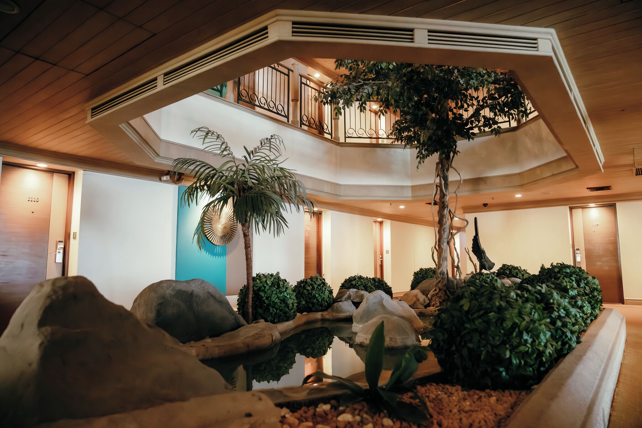 A multi-level atrium with a central water feature, surrounded by various plants and a large tree.