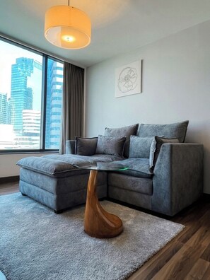 A modern living room with a grey sectional sofa, a wooden coffee table, and a city view through large windows. - Awtan Sukhumvit (Bangkok)