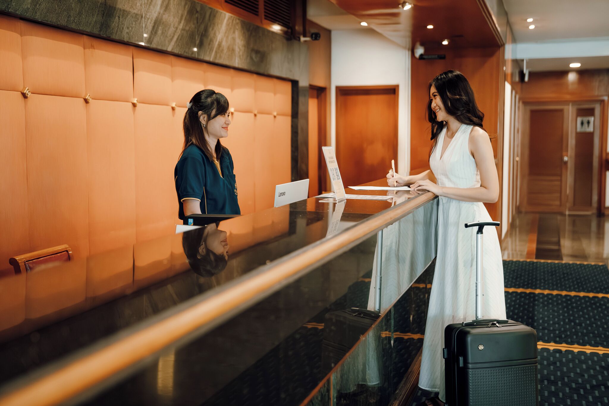 Two individuals at a hotel reception desk with a luggage trolley and a Lenovo sign.