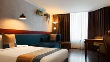 A hotel room with a large bed, a sofa, a desk, and a chair. There are two wall-mounted lamps and a window with curtains.