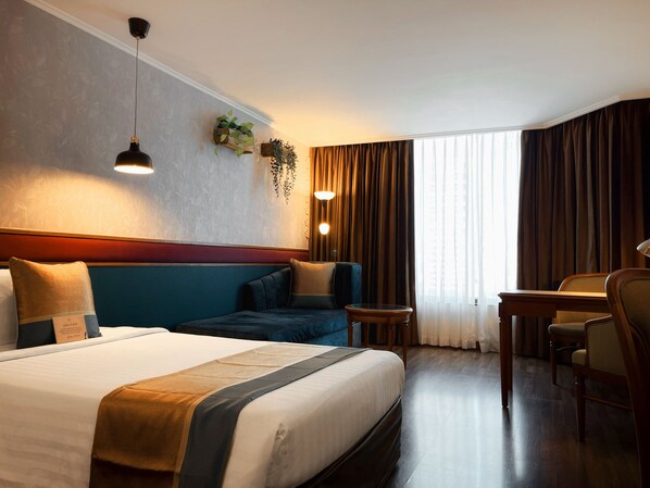 A hotel room with a large bed, a sofa, a desk, and a chair. There are two wall-mounted lamps and a window with curtains. - Awtan Sukhumvit (Bangkok)