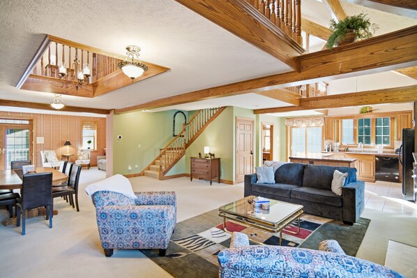 A spacious living area with a staircase, wooden beams, a dining set, and a kitchen in the background.