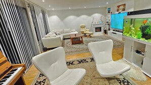 A modern living room with a piano, white chairs, a TV, and a fish tank. - Spacious Bergen House With Stunning Mountain Views (Bergen)