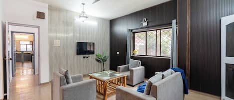 A modern living room with grey sofas, a glass coffee table, and a flat-screen TV mounted on the wall.