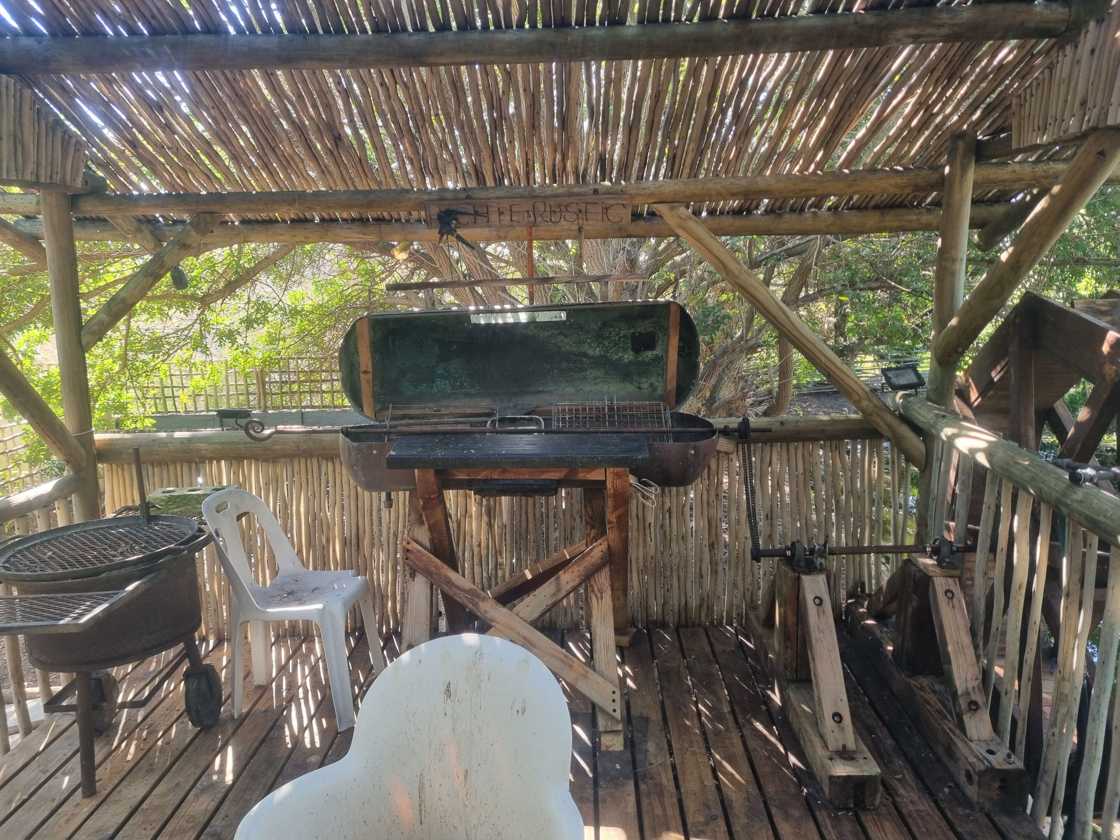 A rustic outdoor grill area with a wooden structure, a metal grill, and a white plastic chair.