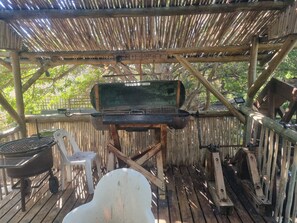 Ein rustikaler Grillbereich im Freien mit einer Holzkonstruktion, einem Metallgrill und einem weißen Kunststoffstuhl.