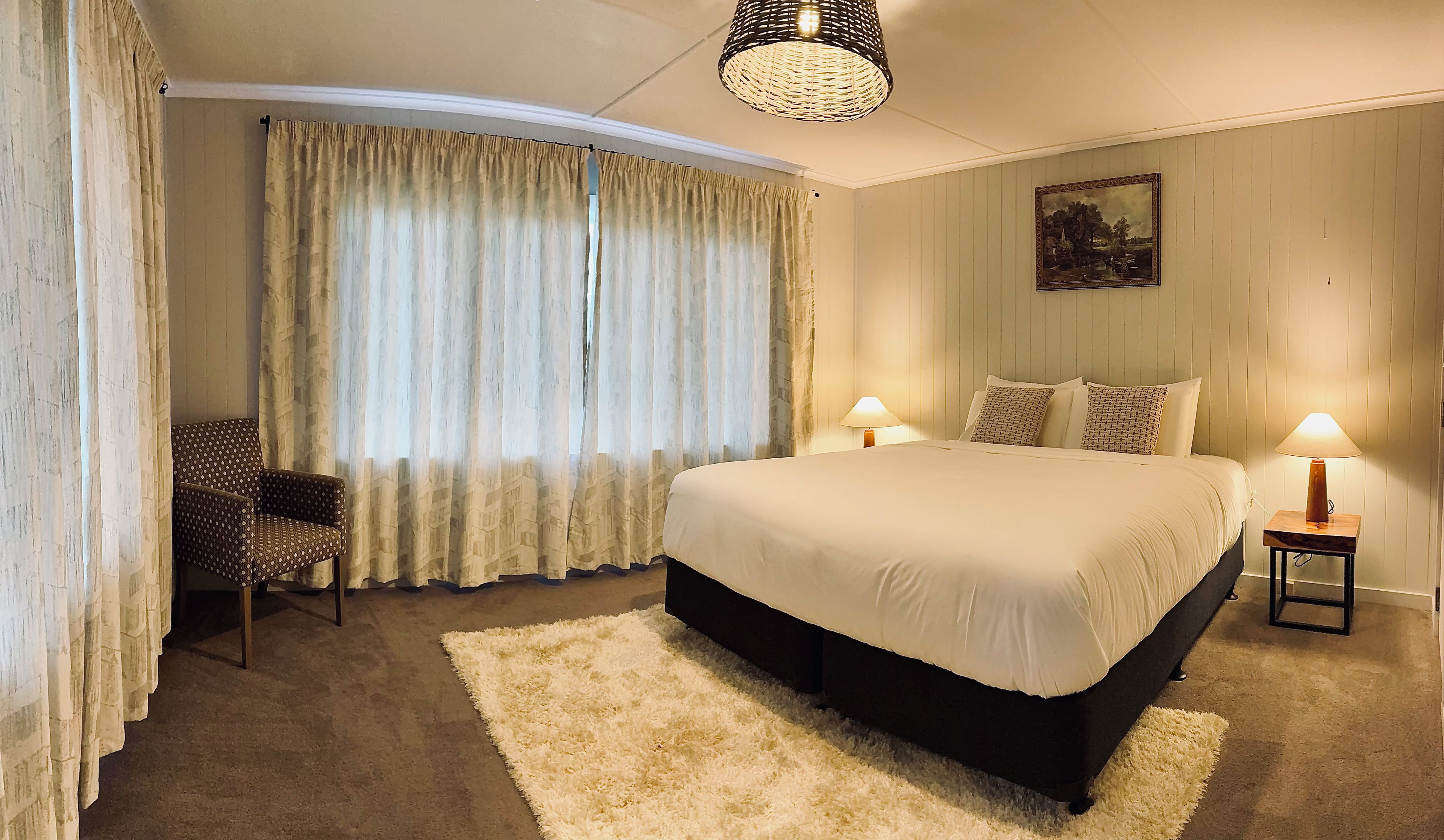 A hotel room with a large bed, two bedside lamps, a chair, a fluffy rug, and a painting on the wall.
