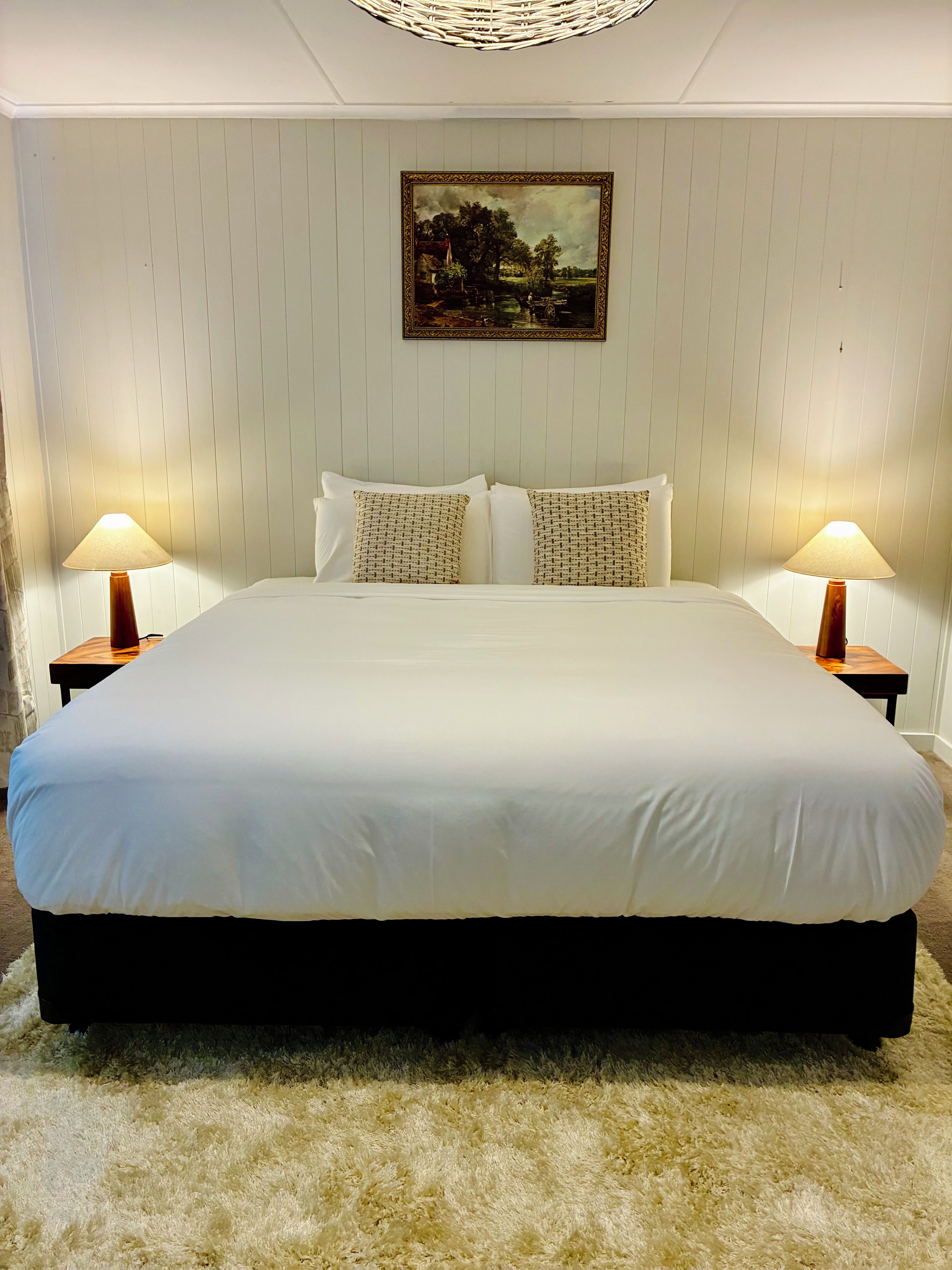 A large bed with white linens, two bedside lamps, and a framed landscape painting on the wall.