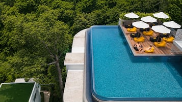 Vue aérienne d’une piscine à débordement sur un toit, avec une terrasse en bois et des fauteuils en forme de sac de haricots jaunes, le tout entouré d’une végétation luxuriante.