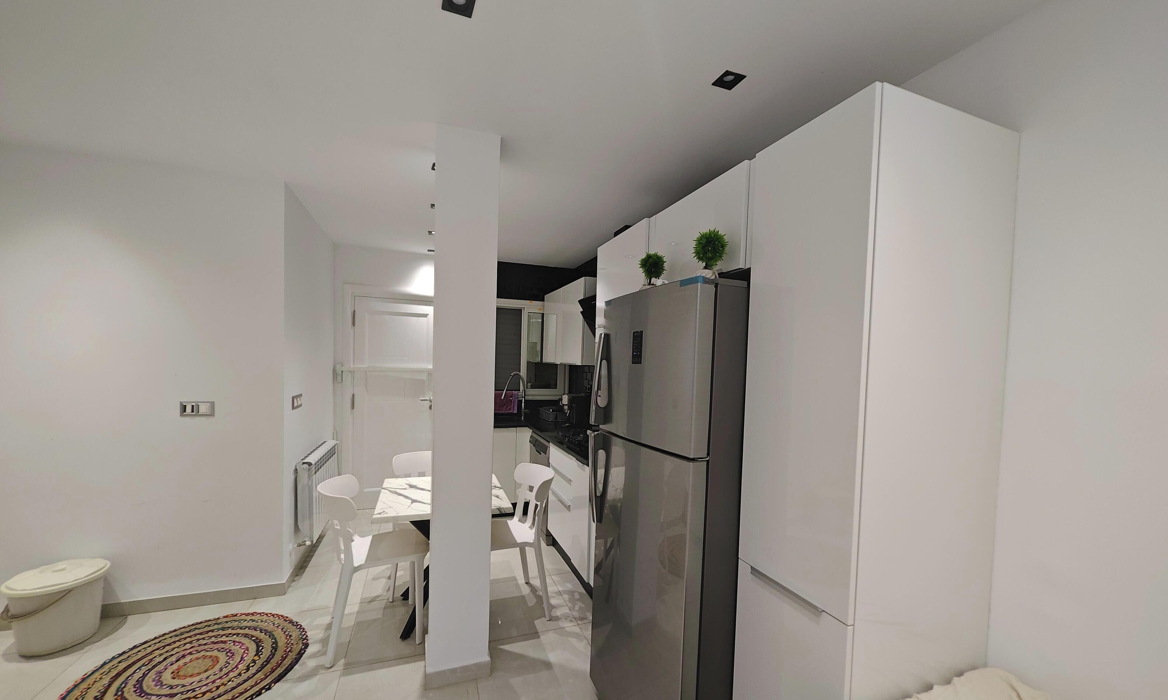 A modern kitchen with white cabinets, a stainless steel refrigerator, and a dining area with a white table and chairs.