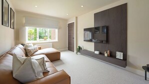 A modern living room with a leather sofa, a flat-screen TV mounted on a wooden panel, and a large window with a view of greenery. - Modern Home in Gleneagles Village | 2 Bed (Auchterarder)