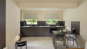 A modern kitchen with dark cabinetry, a central island, and a dining area with metal chairs. - Modern Home in Gleneagles Village | 2 Bed (Auchterarder)