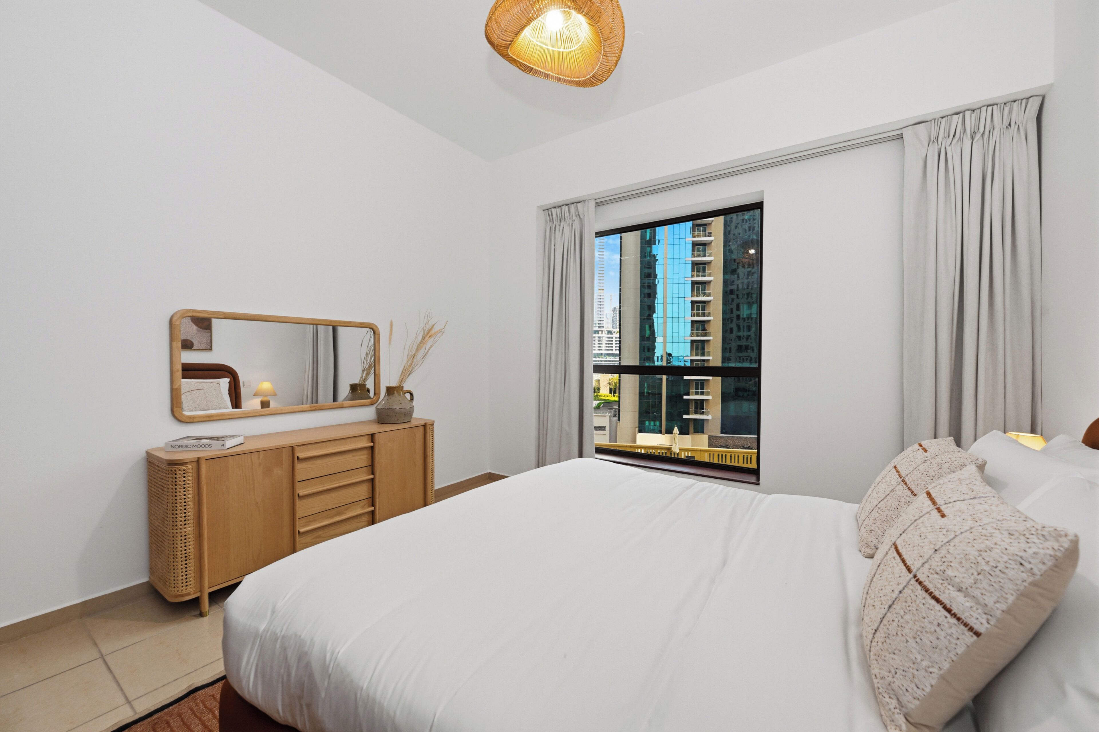 A modern hotel room with a large bed, a wooden dresser, a mirror, and a window with a city view.