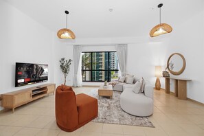 A modern living room with a flat-screen TV, a sofa, a coffee table, and a large window with a view of high-rise buildings. - Bloomfields 3br in Rimal 4 JBR (Dubai)