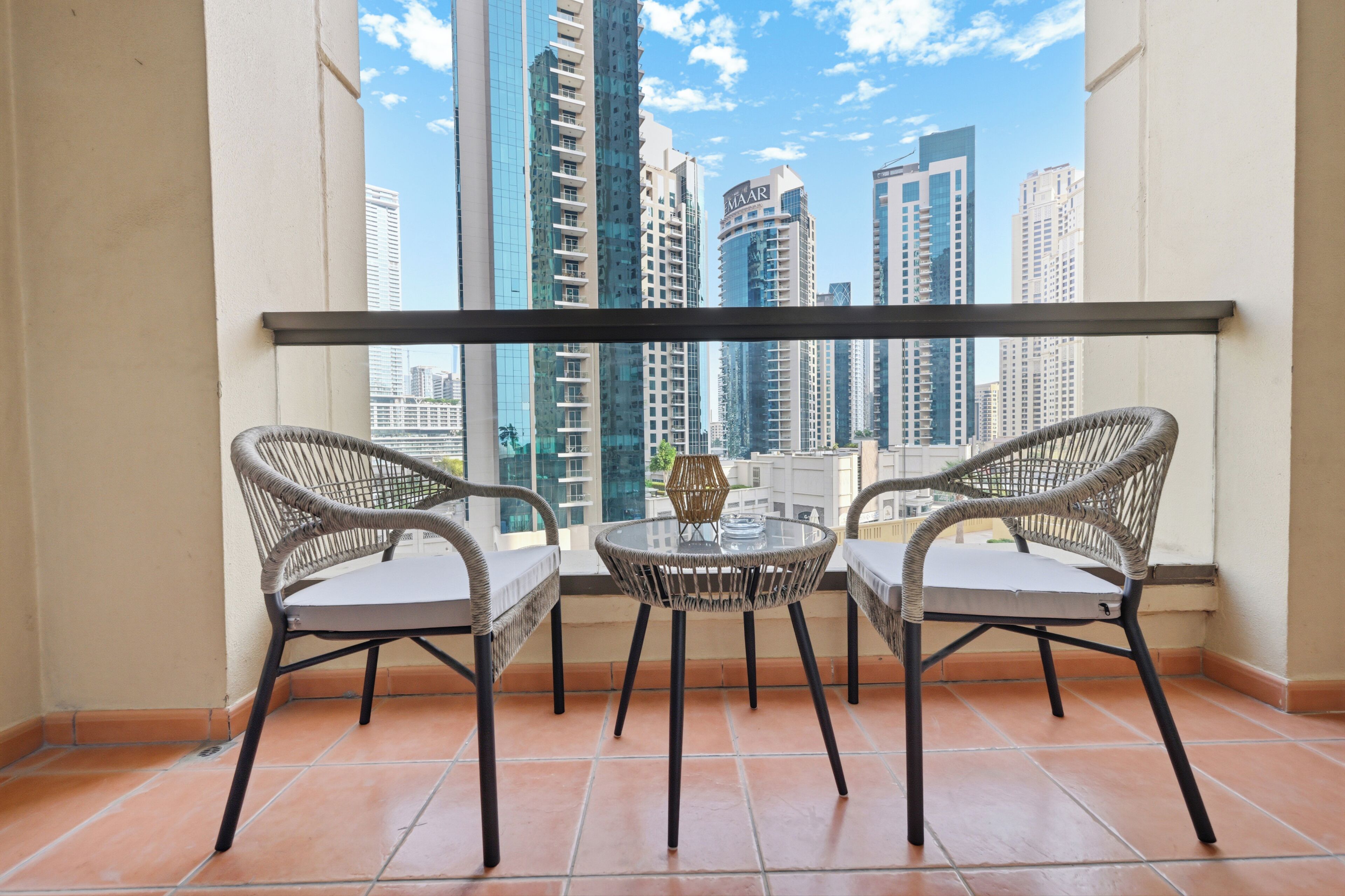 Uma varanda com duas cadeiras de vime e uma pequena mesa redonda, com vista para o horizonte urbano com edifícios altos.