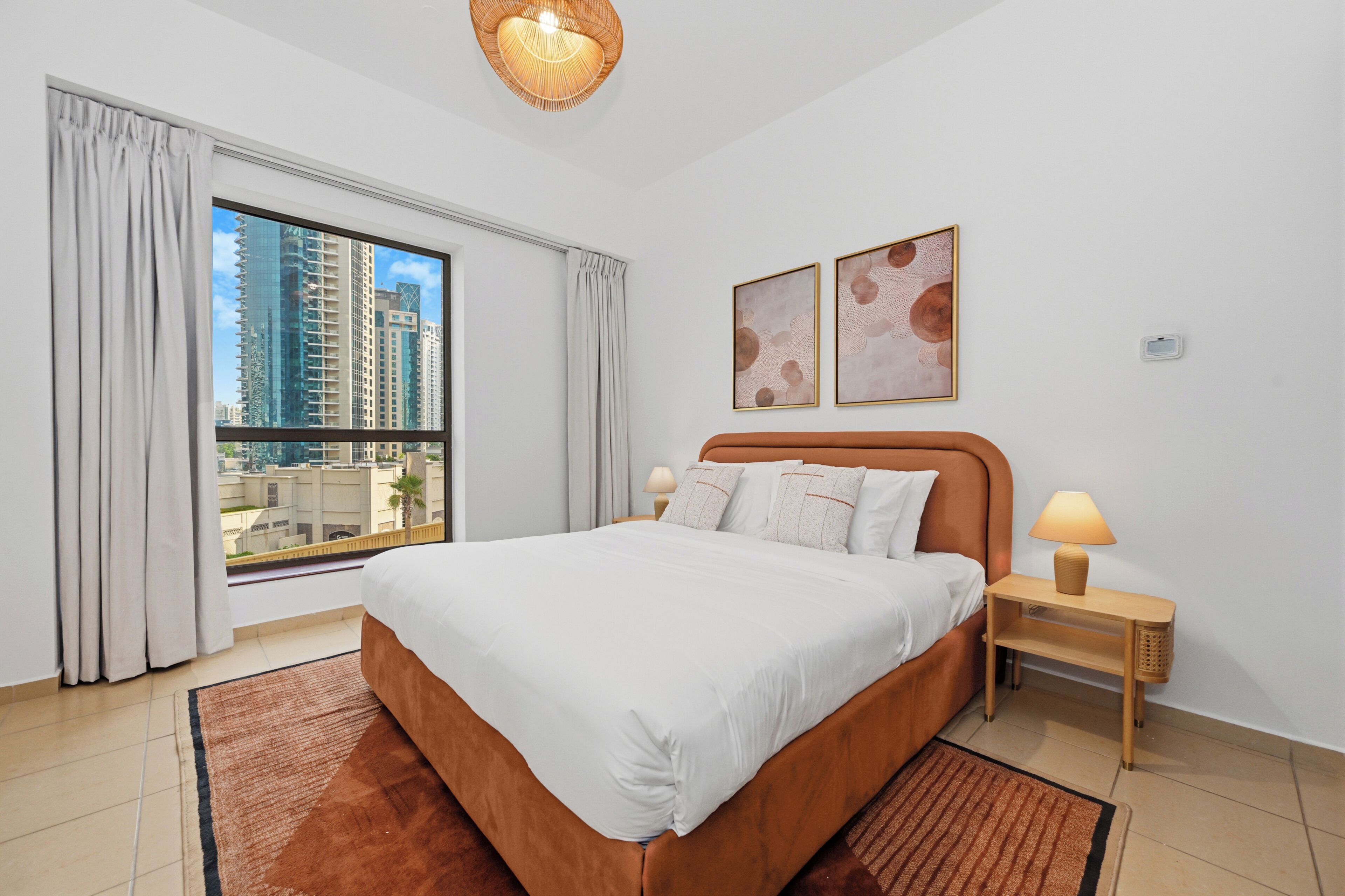 Habitación de hotel con una cama grande, dos lámparas de noche, vistas a edificios altos y dos cuadros en la pared.