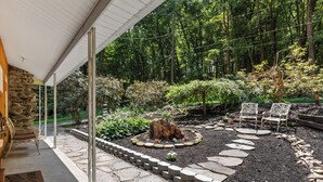 A stone patio with a fire pit, surrounded by a garden with various plants and trees. There are two chairs and a table under a covered area. - Lakefront Retreat,Sleeps 8, Kayaks & Fishing (Pine Grove)