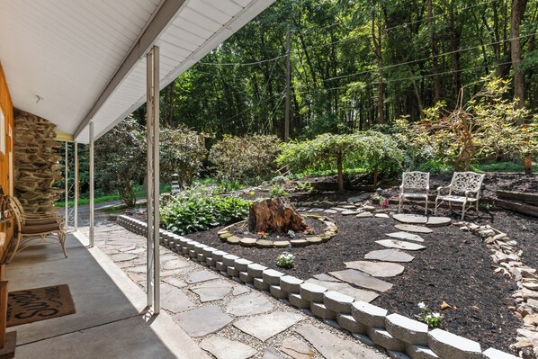 A stone patio with a fire pit, surrounded by a garden with various plants and trees. There are two chairs and a table under a covered area.