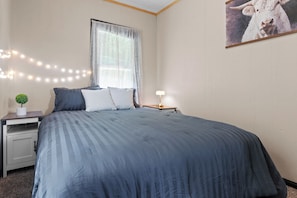A bedroom with a large bed, a nightstand with a lamp, a small plant, and a window with sheer curtains. - Lakefront Retreat,Sleeps 8, Kayaks & Fishing (Pine Grove)