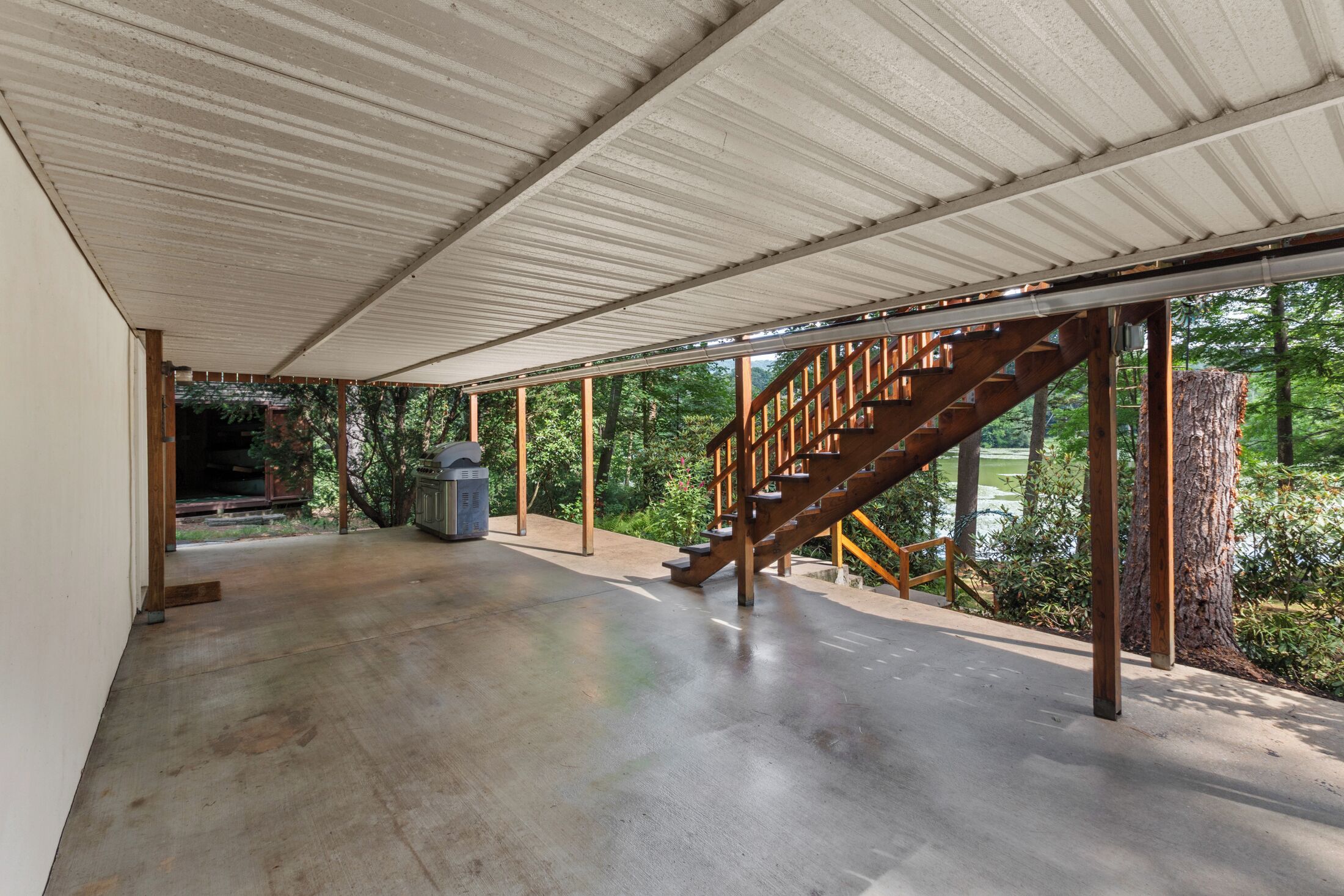 Ett takförsett utomhusområde med ett betonggolv, en trätrappa och utsikt över träd och en vattenyta.