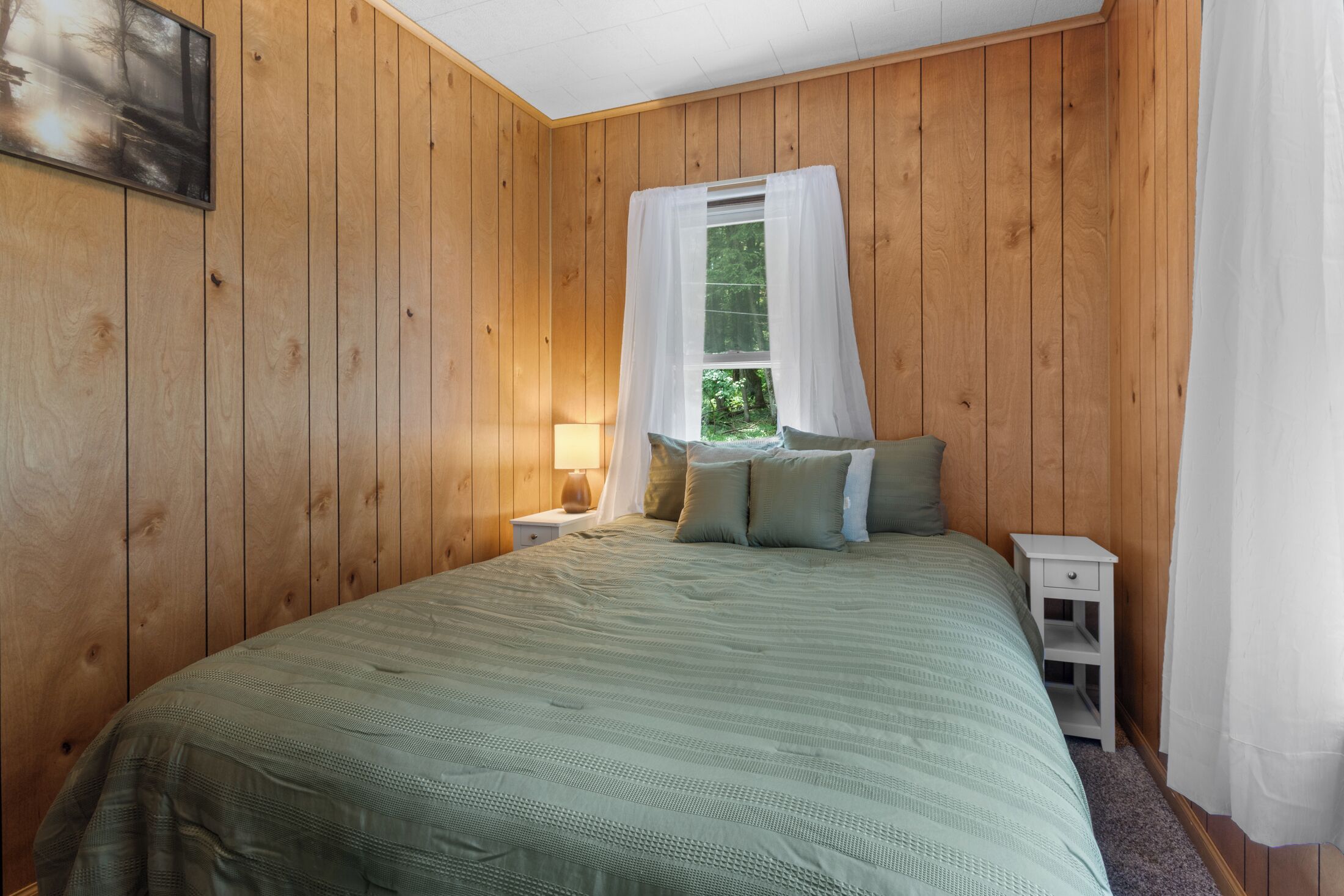 A room with wooden panel walls, a bed with green bedding, a nightstand with a lamp, and a window with white curtains.