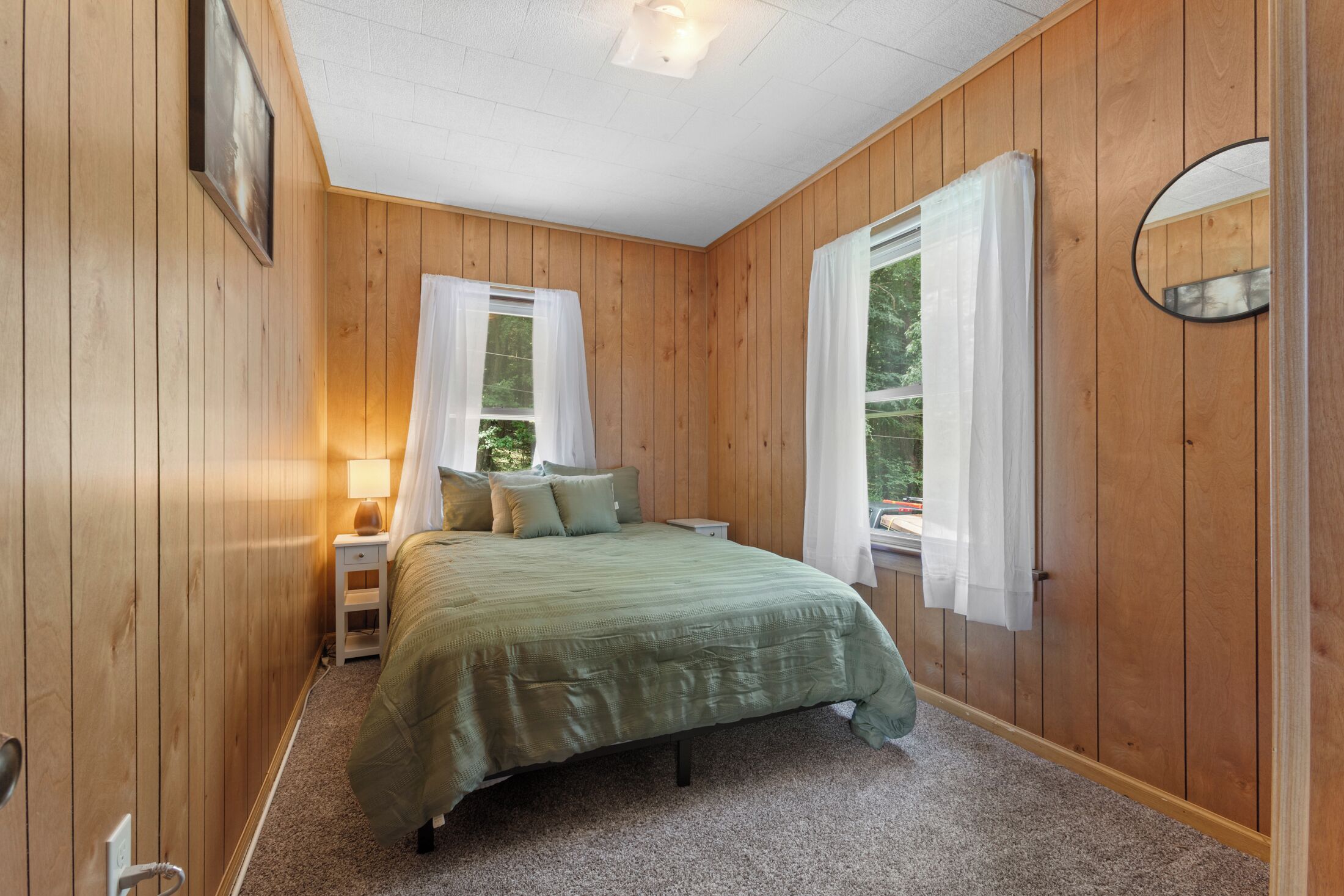 Una camera da letto con pareti in legno, un letto con copripiumino verde, due finestre con tende bianche, un comodino con una lampada e uno specchio appeso al muro.