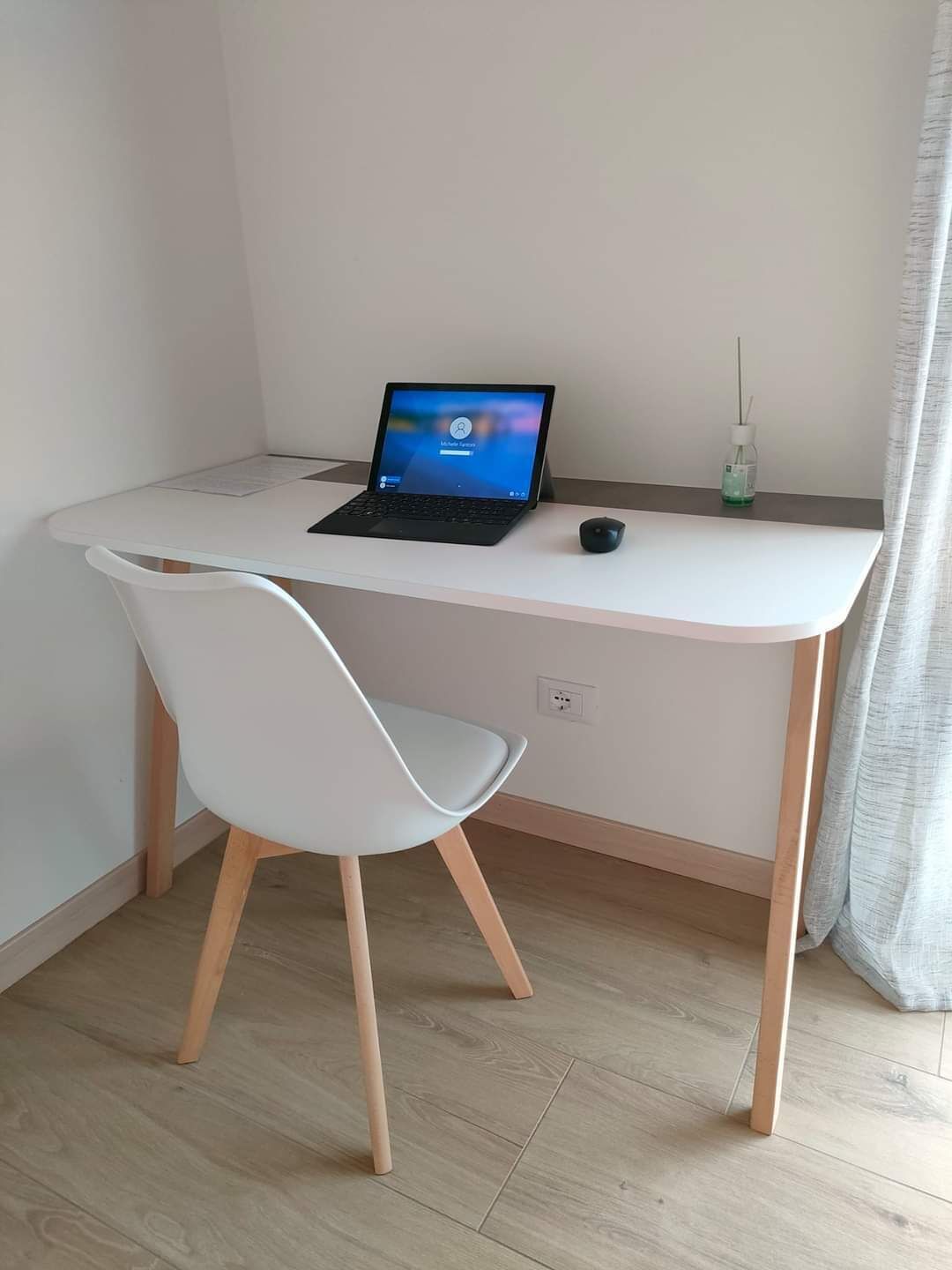 A small, modern room with a white desk, a laptop, a chair, and a small table with a bottle.