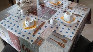 Een eettafel gedekt met witte kopjes, schoteltjes en een broodmand, met een tafelkleed met stippen en een bord waarop staat 'LOVE COOKING'.