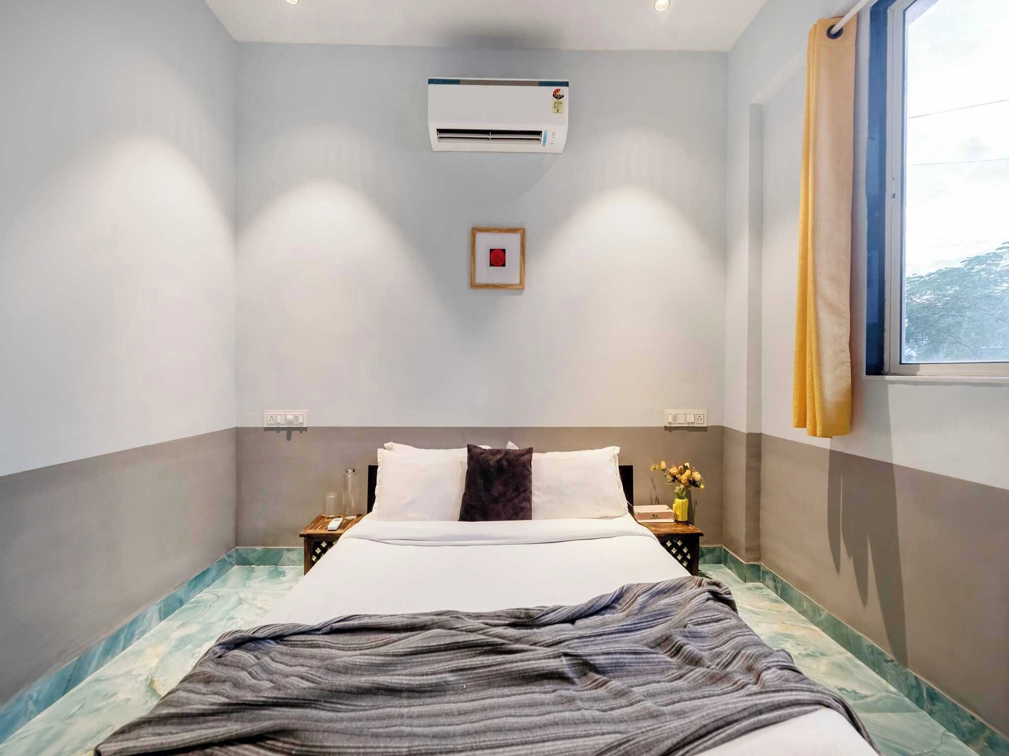 A hotel room with a bed, an air conditioning unit, a nightstand, a bedside table with a lamp, and a window with curtains.
