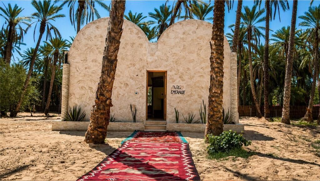 Un edificio con una entrada arqueada distintiva, palmeras y una alfombra roja con motivos que conduce a la puerta.