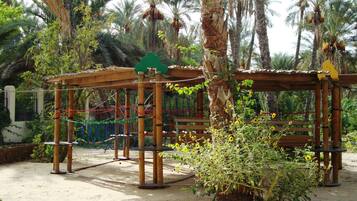 A wooden play structure with ropes and a green sign, surrounded by palm trees and plants.