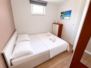 A neatly made bed with pillows and a blanket, a small bedside table with a lamp, a window with a view of the beach, and a painting on the wall. - Spacious 3-bedroom apartment in gorgeous Podstrana (Podstrana - Sv. Martin)