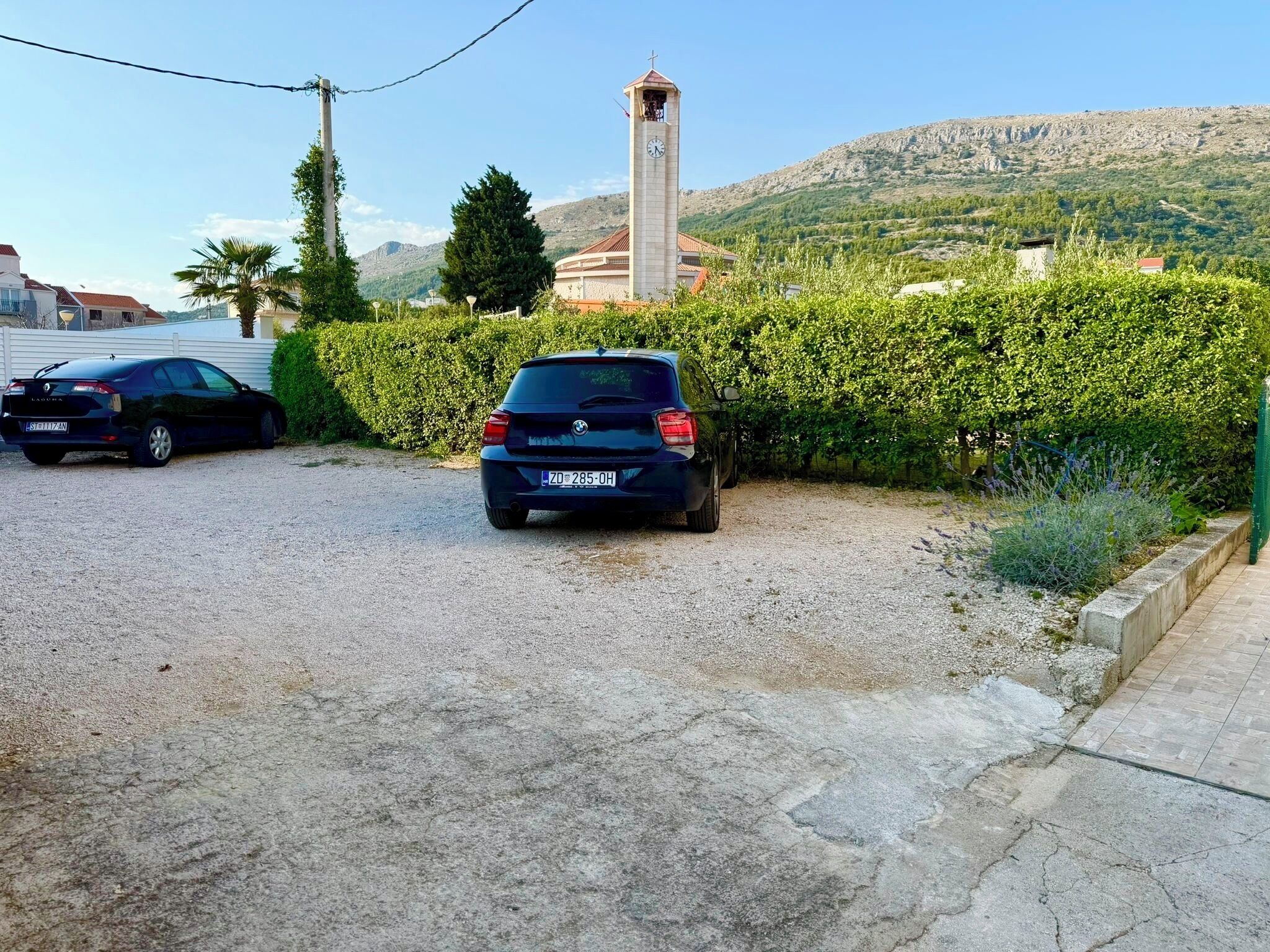 A gravel parking area with two cars parked, a church with a clock tower, and a mountainous landscape in the background.