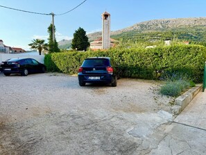 A gravel parking area with two cars parked, a church with a clock tower, and a mountainous landscape in the background. - Spacious 3-bedroom apartment in gorgeous Podstrana (Podstrana - Sv. Martin)