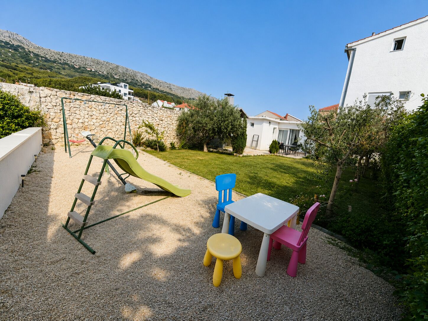 Ein Kinderspielbereich mit Rutsche, Tisch und Stühlen, umgeben von einer Steinmauer und viel Grün.