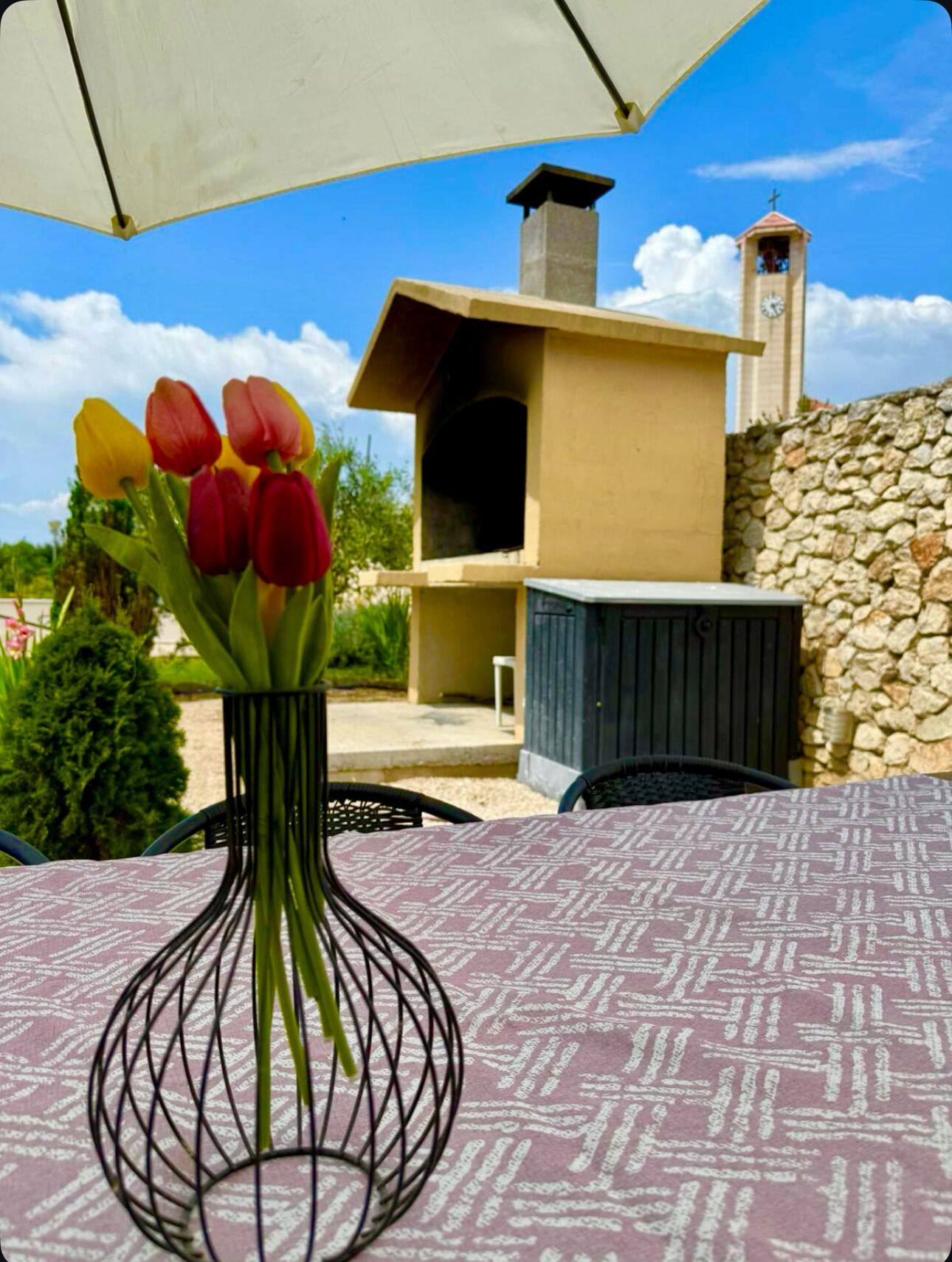 A table with a patterned tablecloth, a vase with tulips, and a stone wall with a chimney and a clock tower in the background.