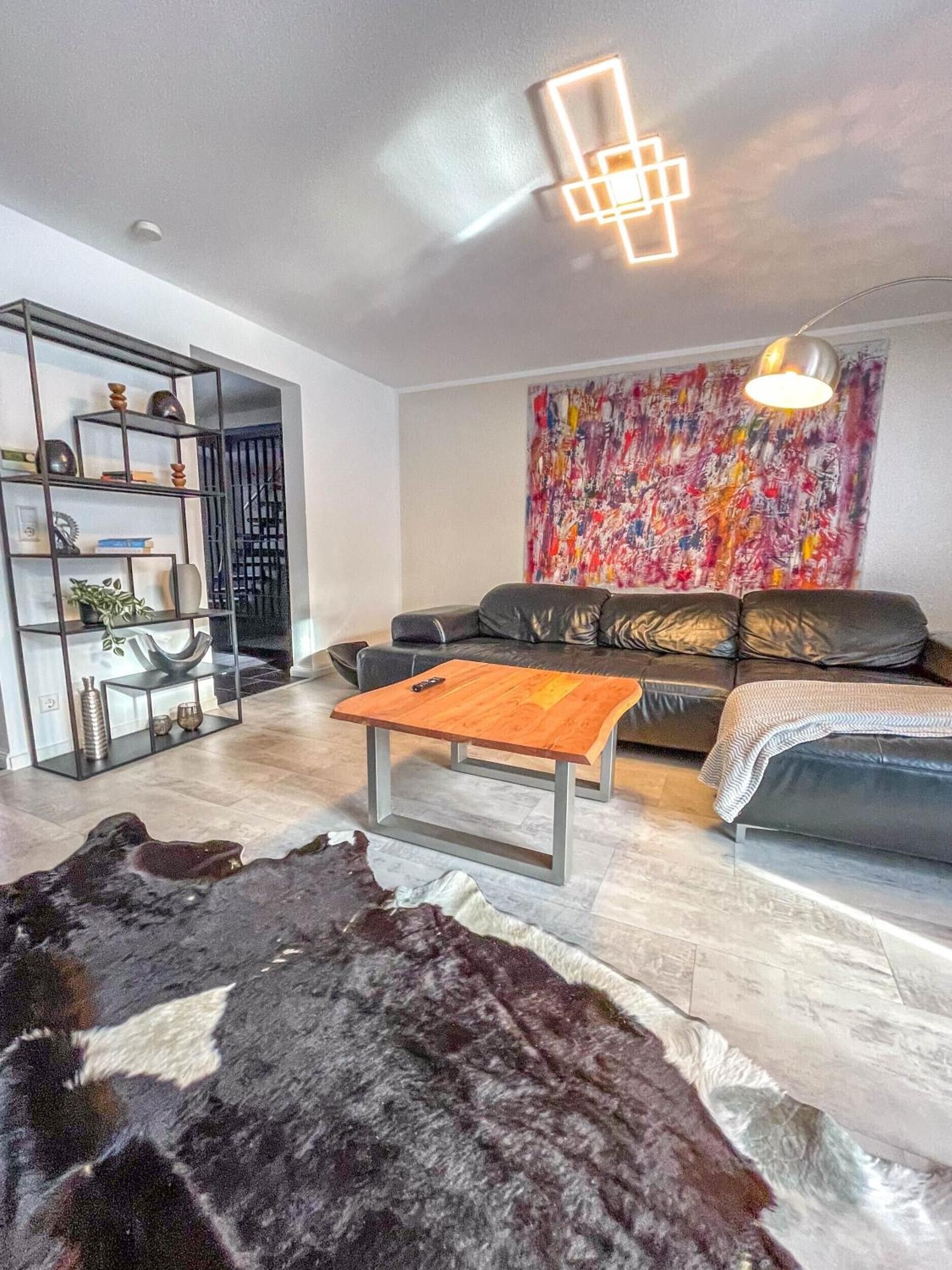 A modern living room with a leather sofa, a wooden coffee table, a black and white rug, a black metal shelving unit, and a colorful abstract painting on the wall.