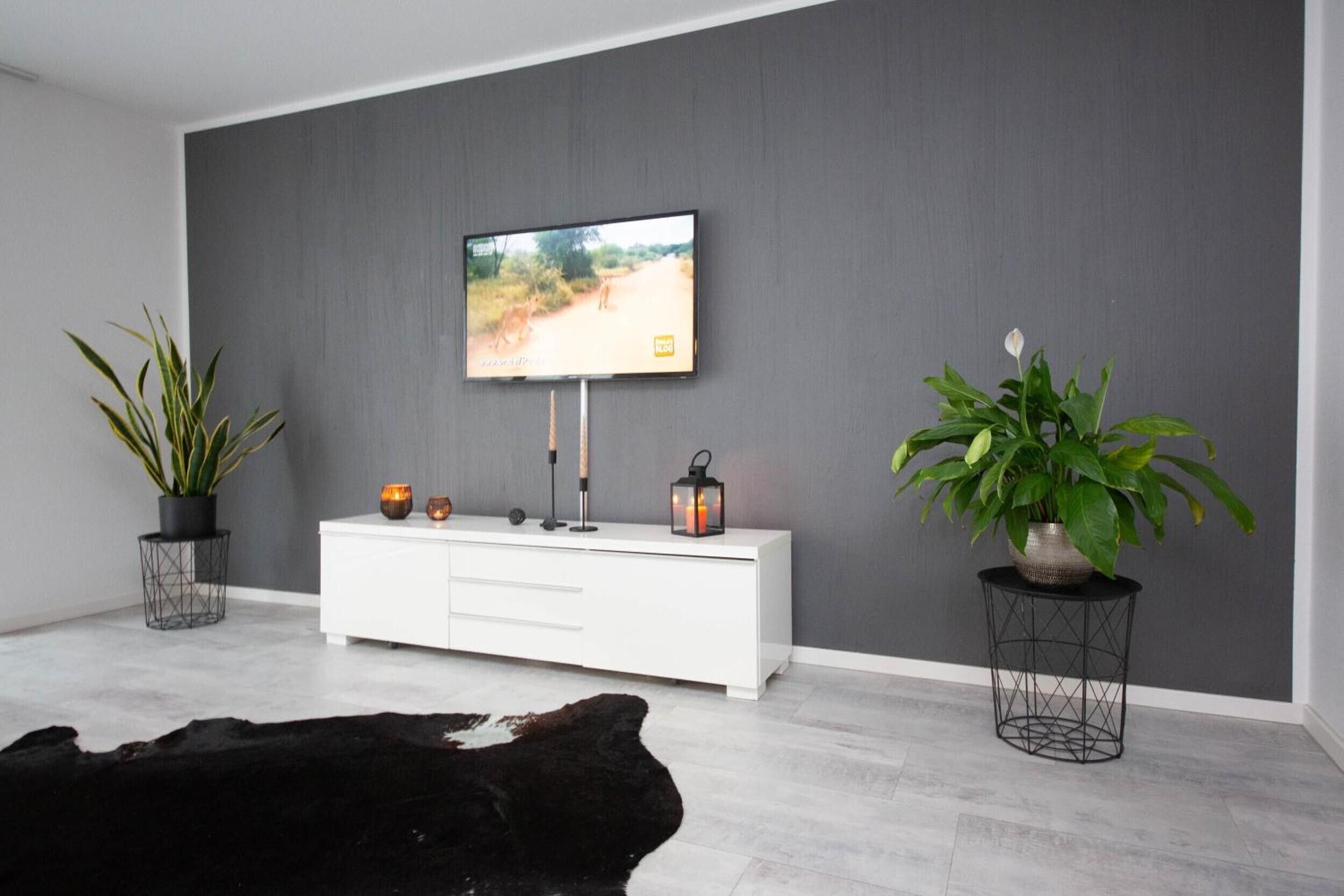 A modern living room with a grey wall, a mounted TV, a white cabinet, two potted plants, and a black leather sofa.