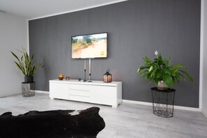A modern living room with a grey wall, a mounted TV, a white cabinet, two potted plants, and a black leather sofa.
