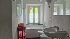 Ein Badezimmer mit einer Badewanne, zwei Waschbecken, einer Toilette und einem Fenster mit Blick auf Grünflächen.