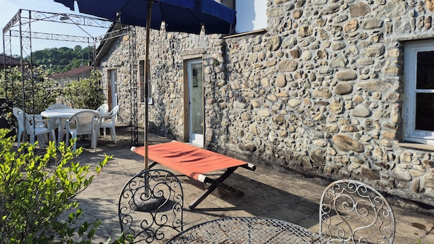 Ein Steinhaus mit einer Terrasse, die Gartenmöbel, einen blauen Sonnenschirm und einen Metallgrill zeigt.