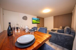 A dining area with a wooden table set for two, a bottle of wine, and a television displaying a landscape.